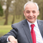 Mike Hedges, wearing a grey suit and red tie, seated on a park bench outdoors in a public green space.