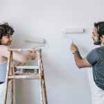 smiling ethnic couple painting wall with roller brushes at home
