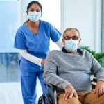 Healthcare worker in scrubs stands behind masked patient in wheelchair, highlighting hospital mask policy during winter virus surge.