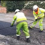Two council workers laying and compacting asphalt during a patch repair on a damaged road surface.