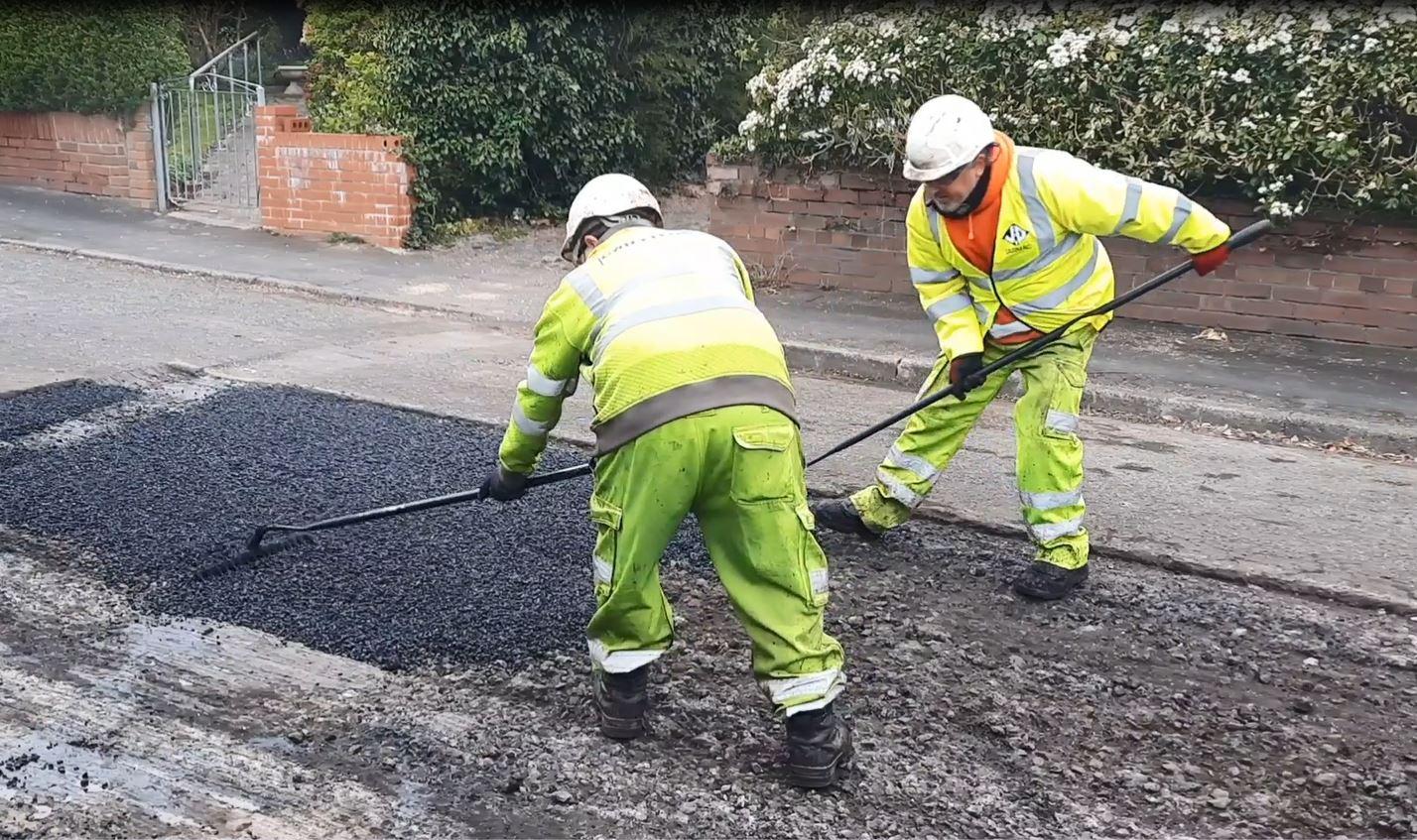 Two council workers laying and compacting asphalt during a patch repair on a damaged road surface.