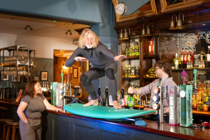 Owen from Surf Therapy, one of the grassroots sports clubs supported by the Greene King IPA Proud To Pitch In campaign, stands on a surfboard on the bar at the Wig and Pen pub in Oxford. (Richard Dawson/PA Media Assignments)