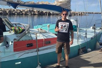 Leigh Evans standing on shore next to the yacht he is joining for the Global Sumud Flotilla aid mission.