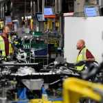 Two Jaguar Land Rover employees working on the Range Rover production line, wearing protective gear and assembling vehicles on the factory floor.