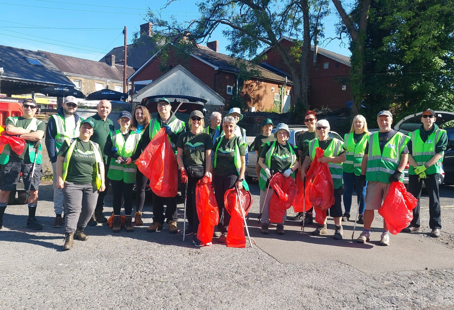 A group of litter‑picking volunteers in Pontardawe wearing green branded T‑shirts and fluorescent vests, holding litter‑picking sticks and red rubbish sacks during a community clean‑up.