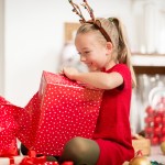 A smiling child sits on the living room floor opening a large red Christmas gift, symbolising the joy of festive family celebrations.