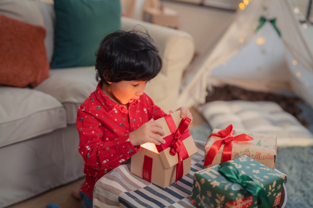 A joyful child opens a present during Christmas, with wrapped gifts and a cosy living room setting in the background.