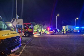 Night‑time scene showing ambulance, fire and police vehicles with blue lights at the B4304 roundabout near Llwynhendy, where crews used specialist equipment to rescue two people after a car left the road.