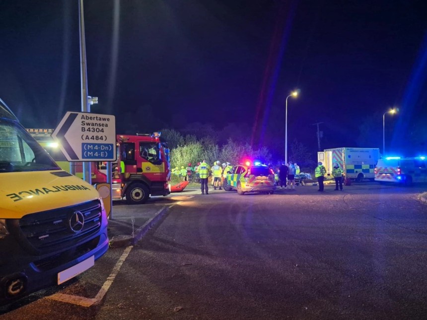 Night‑time scene showing ambulance, fire and police vehicles with blue lights at the B4304 roundabout near Llwynhendy, where crews used specialist equipment to rescue two people after a car left the road.