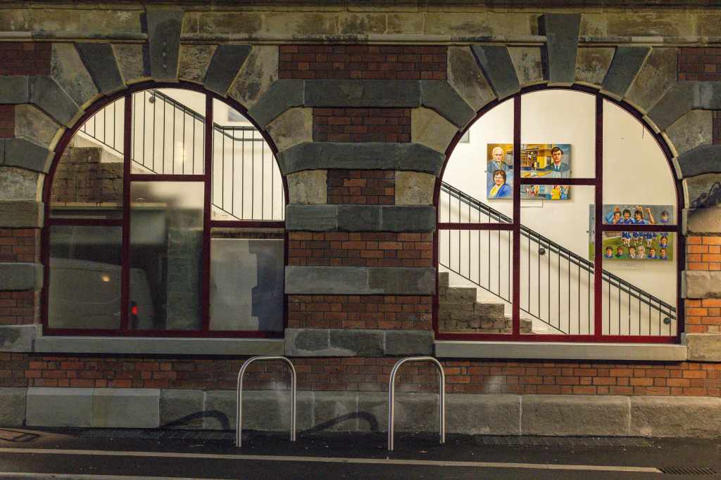 Night view of the side of Swansea’s Palace Theatre showing two illuminated arched windows, interior stairwell artwork, and bicycle stands outside.