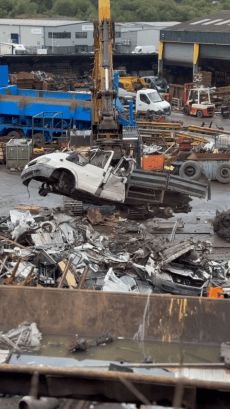 White transit pickup truck being crushed by industrial machinery at a scrapyard, with other vehicles and metal debris visible in the background