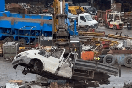 White transit pickup truck being crushed by industrial machinery at a scrapyard, with other vehicles and metal debris visible in the background