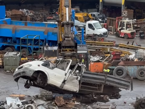 White transit pickup truck being crushed by industrial machinery at a scrapyard, with other vehicles and metal debris visible in the background