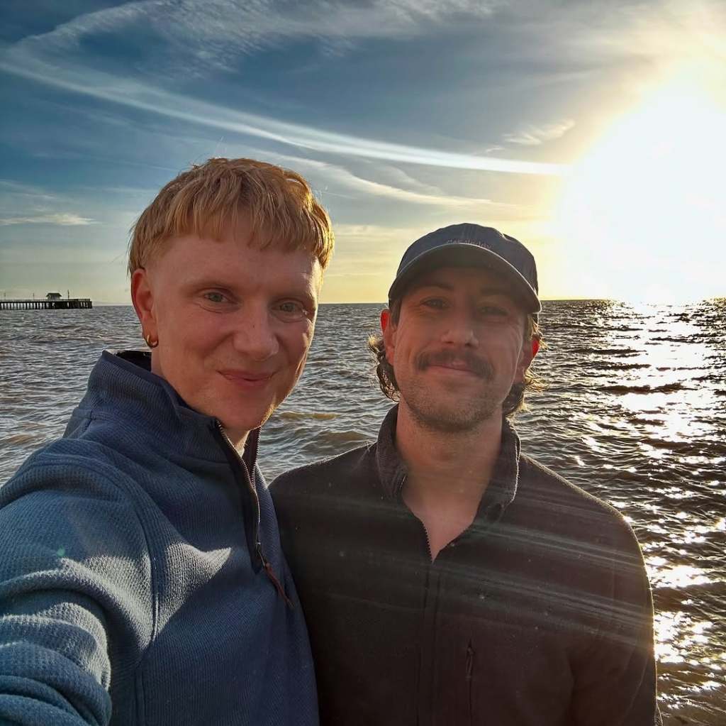 Owain Wyn Evans standing by the water with his husband Arran, posing for the camera.