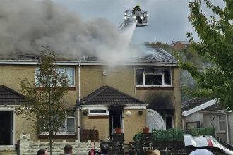 Dramatic images show flames and smoke pouring from the roof of a two‑storey end‑terrace house in Neath as firefighters continue their response.