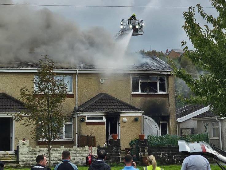 Dramatic images show flames and smoke pouring from the roof of a two‑storey end‑terrace house in Neath as firefighters continue their response.