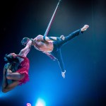 Martin Alvez hangs from a leather strap by one arm while Bethan Williams, in a red dress, grips his neck with her feet and arches back dramatically during a Cirque du Soleil performance.