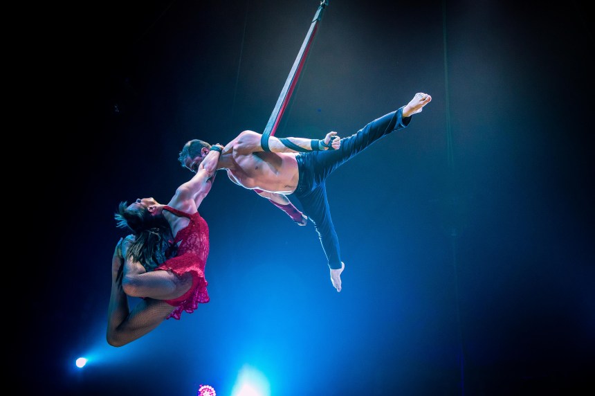 Martin Alvez hangs from a leather strap by one arm while Bethan Williams, in a red dress, grips his neck with her feet and arches back dramatically during a Cirque du Soleil performance.