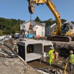 A large excavator lowers a hollow concrete section into a deep trench in Glynneath. Two workers in yellow hi‑vis stand in the trench, one directing the machinery, as part of flood alleviation works.