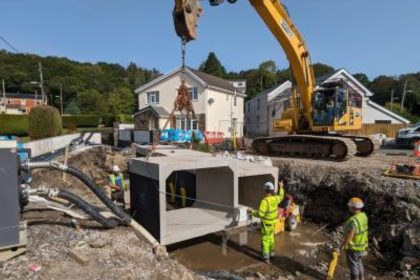 A large excavator lowers a hollow concrete section into a deep trench in Glynneath. Two workers in yellow hi‑vis stand in the trench, one directing the machinery, as part of flood alleviation works.