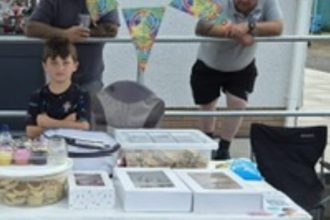 Seven‑year‑old Swansea fundraiser Ollie Blake smiling at his cake stall after cycling 15 miles to raise £1,600 for Faith in Families.