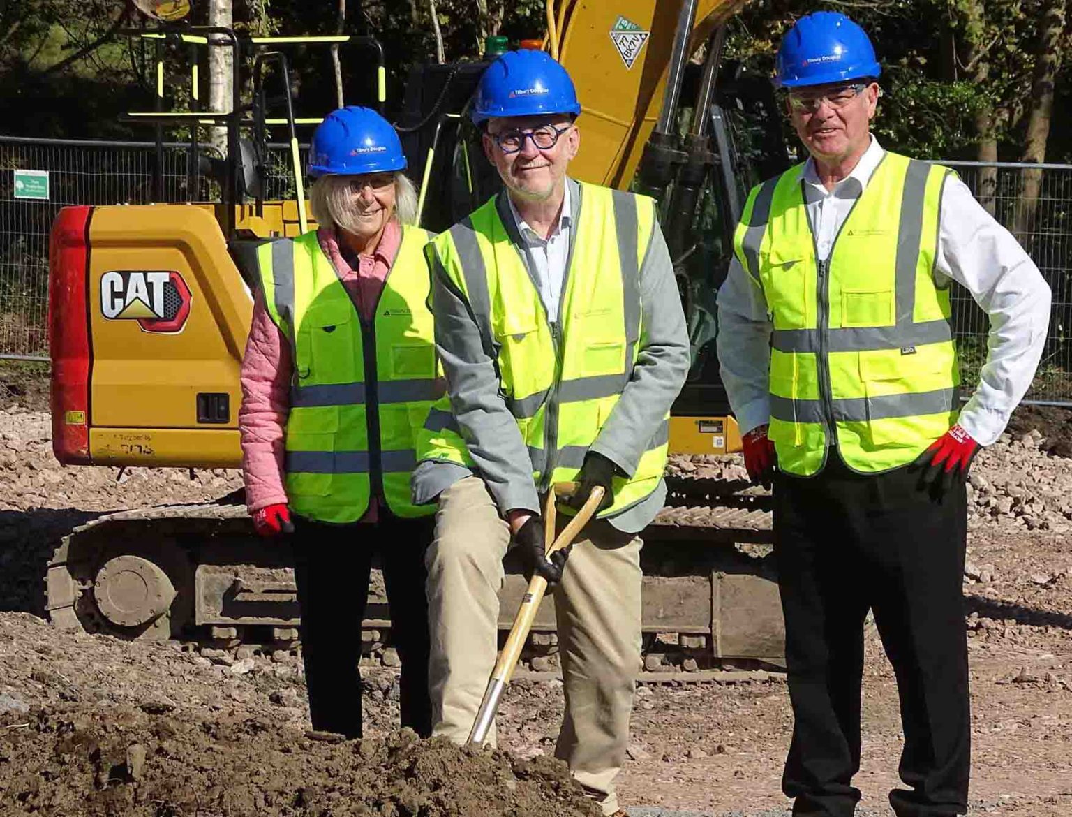 Three people in blue hard hats and yellow high‑visibility vests holding a spade at a ceremonial ground‑breaking in front of a CAT excavator.