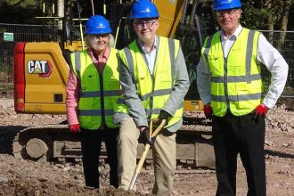 Three people in blue hard hats and yellow high‑visibility vests holding a spade at a ceremonial ground‑breaking in front of a CAT excavator.