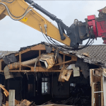 Screenshot shows an excavator with its ripper arm lifted in the air, poised to continue tearing down Cwmfelin Social Club in Cwmbwrla, Swansea.