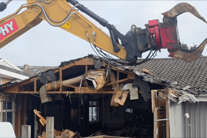 Screenshot shows an excavator with its ripper arm lifted in the air, poised to continue tearing down Cwmfelin Social Club in Cwmbwrla, Swansea.