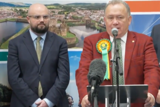 Screenshot from ITV News coverage of the Caerphilly by‑election showing Plaid Cymru’s winning candidate at the podium and Reform UK’s candidate beside him looking disappointed.