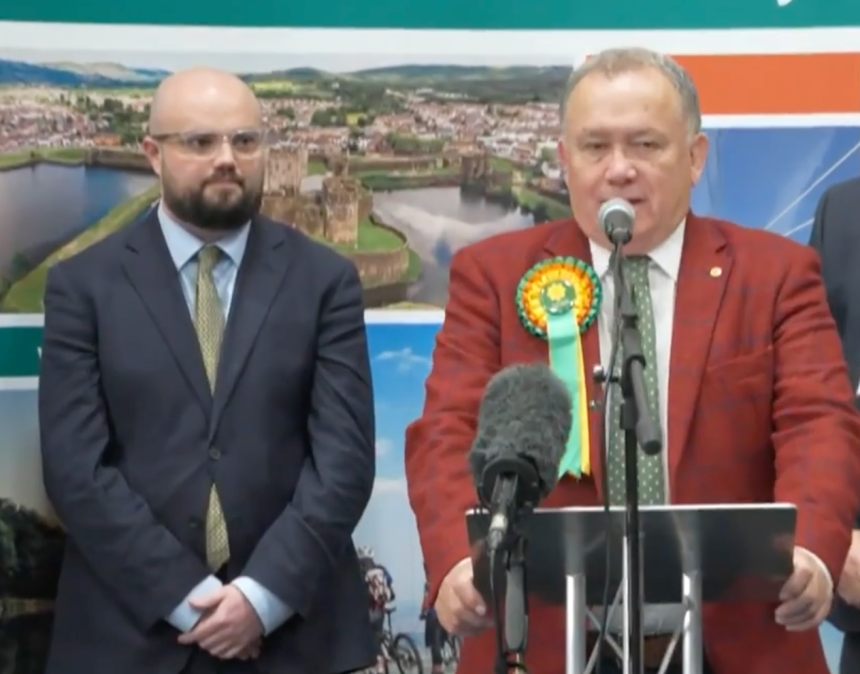 Screenshot from ITV News coverage of the Caerphilly by‑election showing Plaid Cymru’s winning candidate at the podium and Reform UK’s candidate beside him looking disappointed.