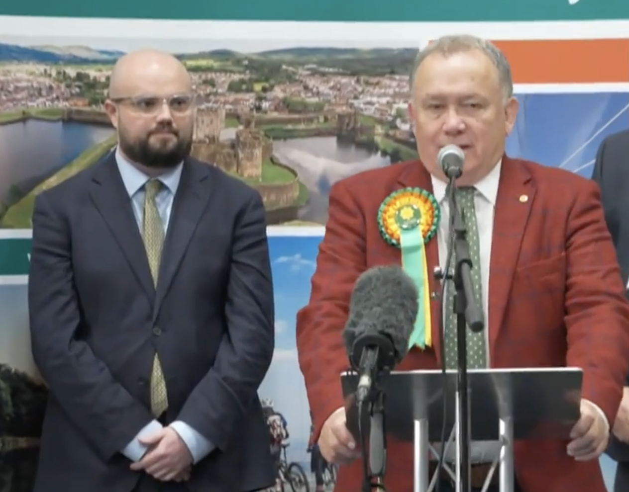 Screenshot from ITV News coverage of the Caerphilly by‑election showing Plaid Cymru’s winning candidate at the podium and Reform UK’s candidate beside him looking disappointed.