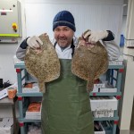 Scott Quinnell smiling while holding two large flatfish during a night shift at Swansea Fish for new S4C series.