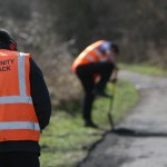 Three people in orange hi‑vis vests on a Community Payback scheme. One, with “Community Payback” written on his back and wearing ear protectors, faces away from the camera. Another digs at the side of a footpath while a probation worker looks on holding a clipboard.