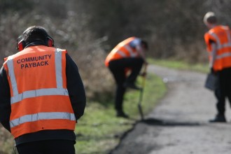 Three people in orange hi‑vis vests on a Community Payback scheme. One, with “Community Payback” written on his back and wearing ear protectors, faces away from the camera. Another digs at the side of a footpath while a probation worker looks on holding a clipboard.