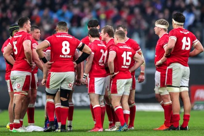 Wales rugby players gathered in a team huddle on the pitch, backs facing the camera.