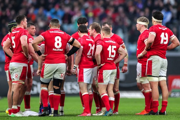 Wales rugby players gathered in a team huddle on the pitch, backs facing the camera.