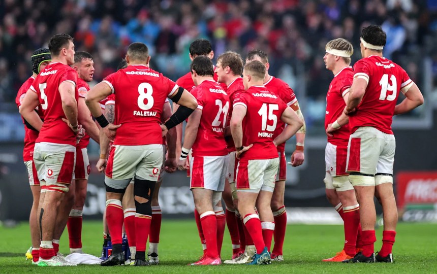 Wales rugby players gathered in a team huddle on the pitch, backs facing the camera.