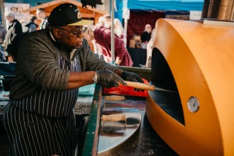 An older food trader works side‑on with a large spatula inside a yellow wood‑fired pizza oven at Neath Food and Drink Festival, showcasing the event’s vibrant street food atmosphere.