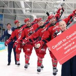 Seven people stand on the ice at Cardiff Devils’ rink, including five players in red jerseys and two officials in business attire, holding a sign promoting new WestJet flights from Cardiff to Toronto. Canadian and Welsh flags are visible in the background.
