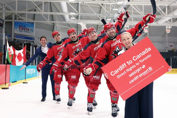 Seven people stand on the ice at Cardiff Devils’ rink, including five players in red jerseys and two officials in business attire, holding a sign promoting new WestJet flights from Cardiff to Toronto. Canadian and Welsh flags are visible in the background.