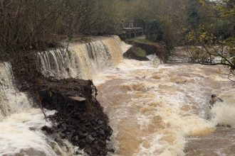 Damage to Kidwelly bridge and dam after flooding, showing collapsed stone sections and fast‑flowing water.