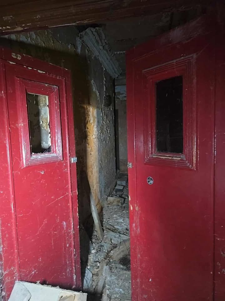 Red double doors opening into a damaged corridor with debris, broken tiles, and peeling walls.