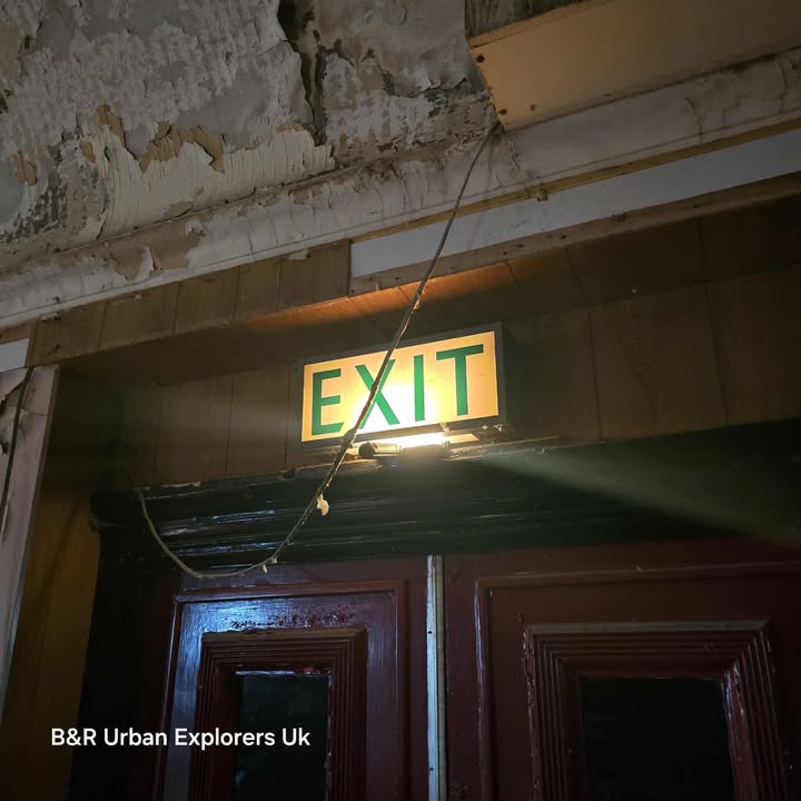 Illuminated exit sign above double doors in a deteriorated interior with peeling paint and hanging wires.