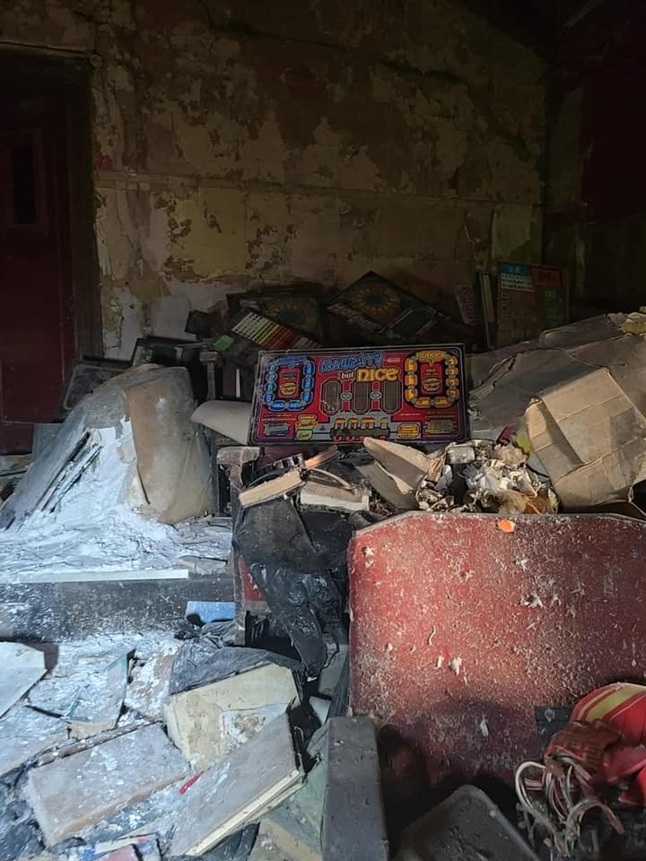 Colourful arcade panel labeled “Naughty but Nice” in abandoned room with broken furniture.