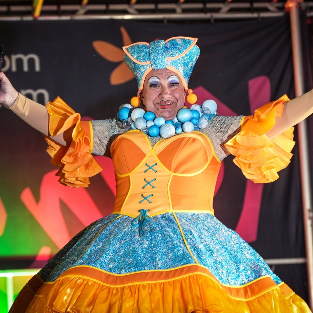 Kev Johns dressed in glittering orange and blue pantomime dame costume with dramatic makeup and headpiece.