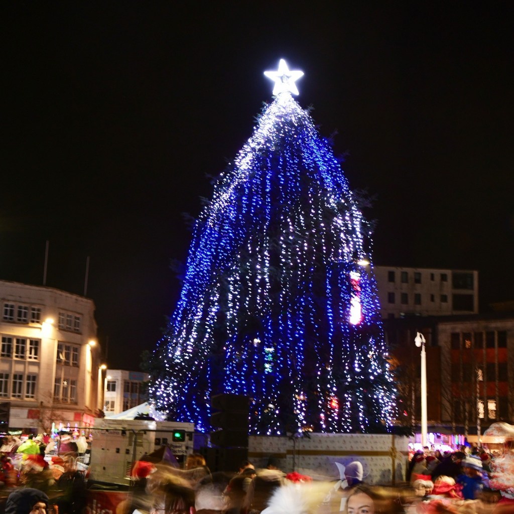 Illuminated Christmas tree with glowing star topper surrounded by festive crowd in city square.