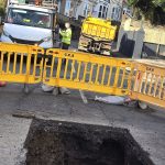 Wide view of Brynymor Road sinkhole site with safety barriers, construction vehicles, and workers.