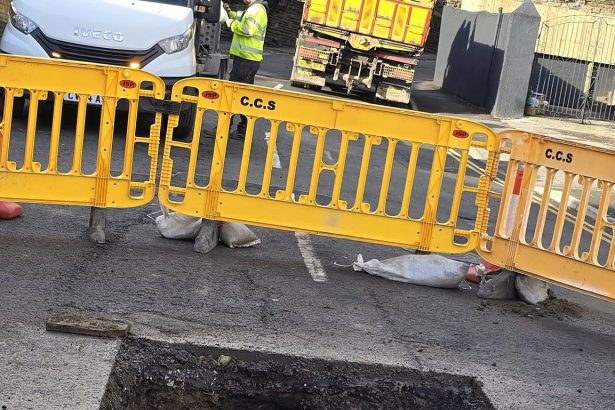 Wide view of Brynymor Road sinkhole site with safety barriers, construction vehicles, and workers.