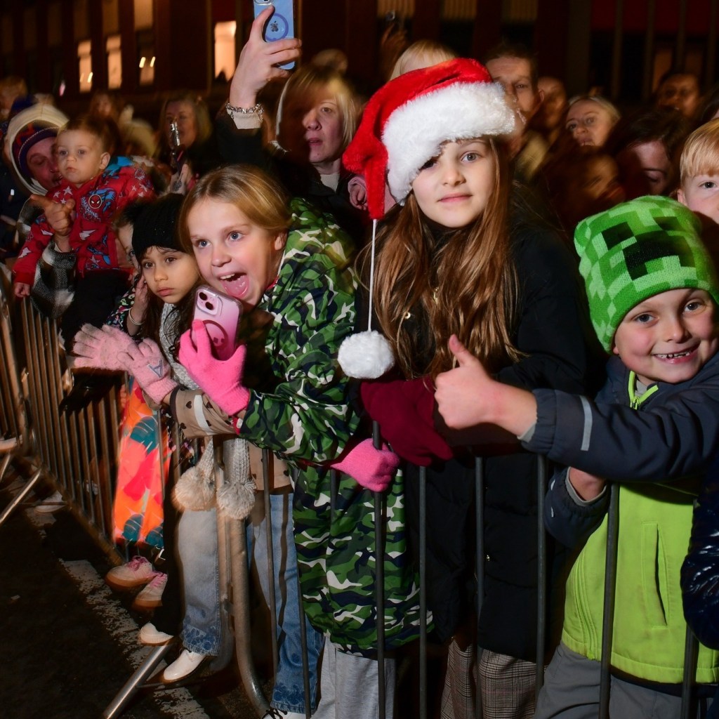 Children in winter clothing behind barricade watching parade, some wearing Santa hats and filming.